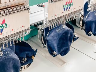 Embroidery machine stitching designs onto multiple blue baseball caps, white and green machine.