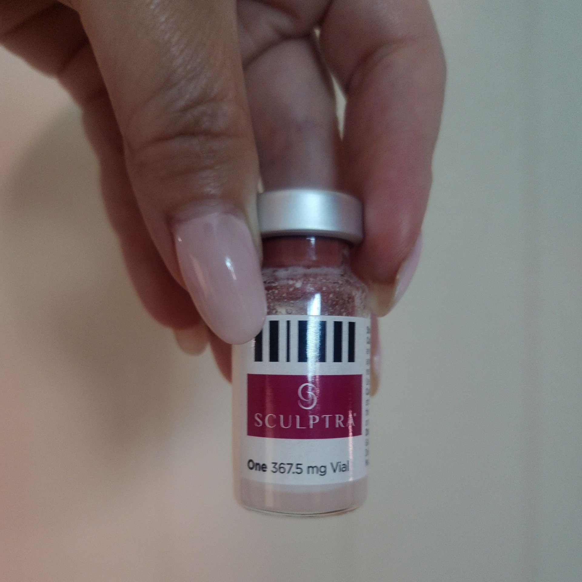 Hand holding a vial of Sculptra, a cosmetic injectable. Bottle is pink with a white label, held against a light background.