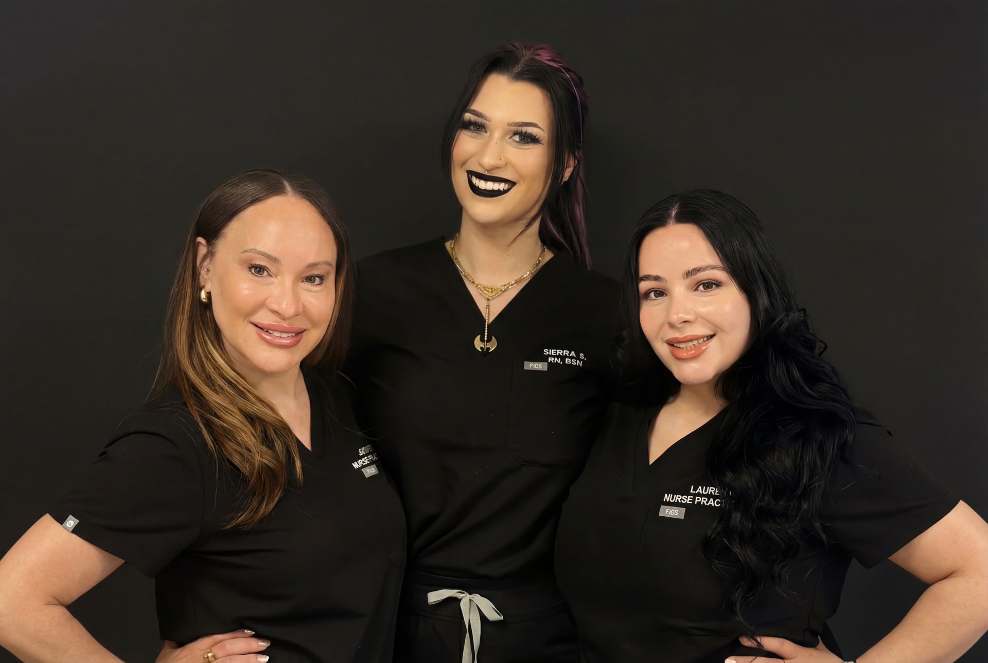 Three people wearing black scrub tops pose together against a black background, smiling and facing the camera.