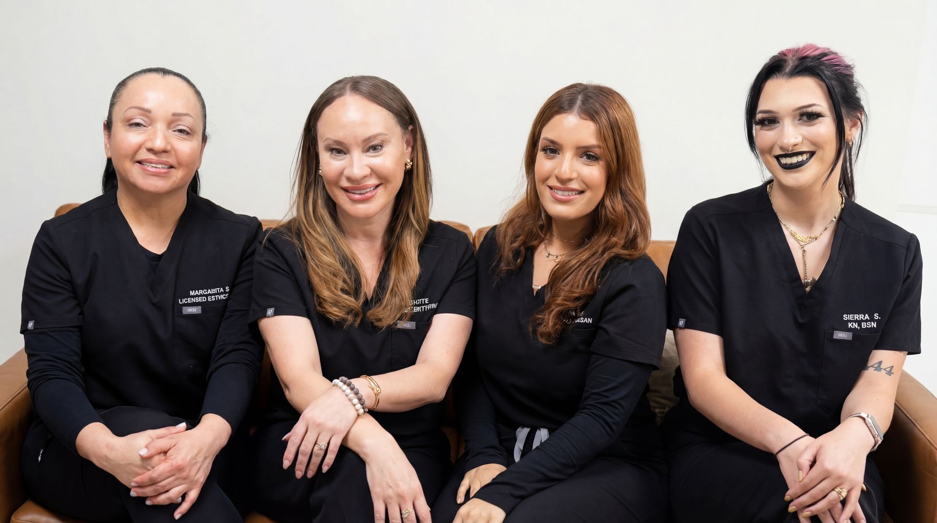 Four individuals in matching black scrub tops sit together on a leather sofa, smiling at the camera against a white wall.