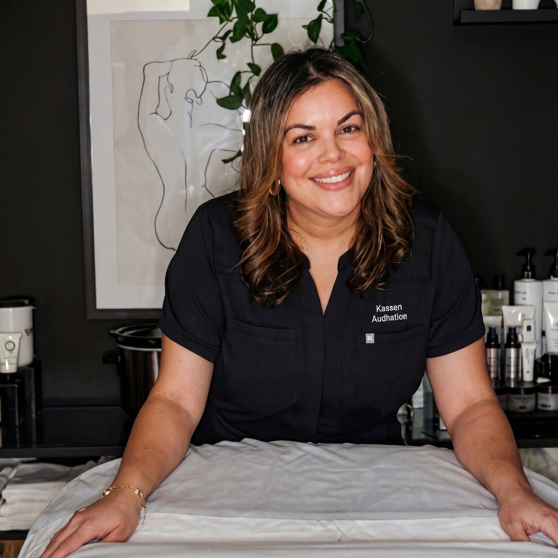 A smiling professional in a dark scrub top standing behind a treatment table in a clinical, neutrally decorated office.