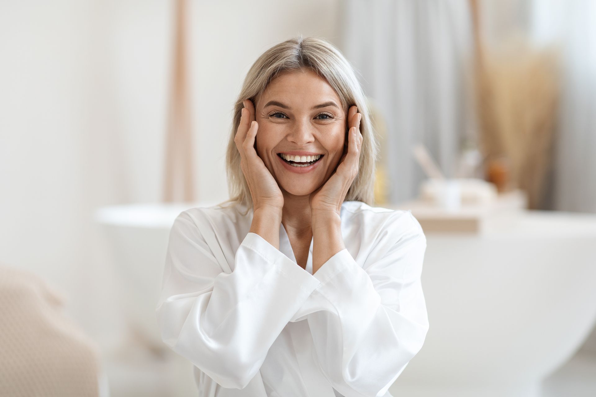 A woman in a bathrobe is smiling and touching her face in a bathroom.