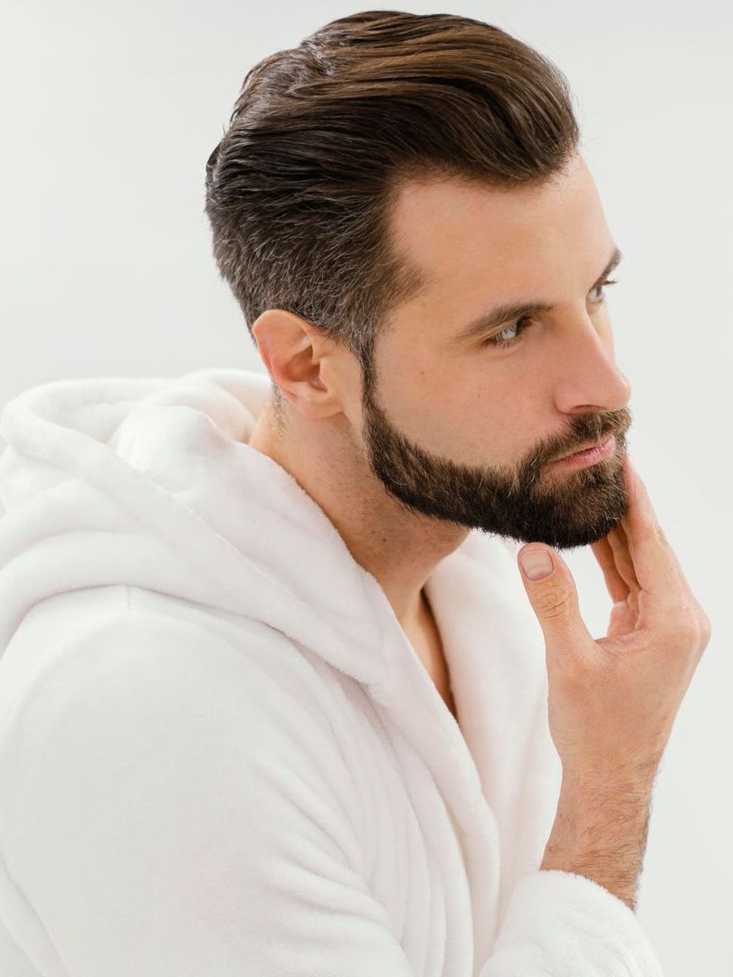 Man in white robe, dark hair with a neatly trimmed beard, gazing thoughtfully, touching his cheek.