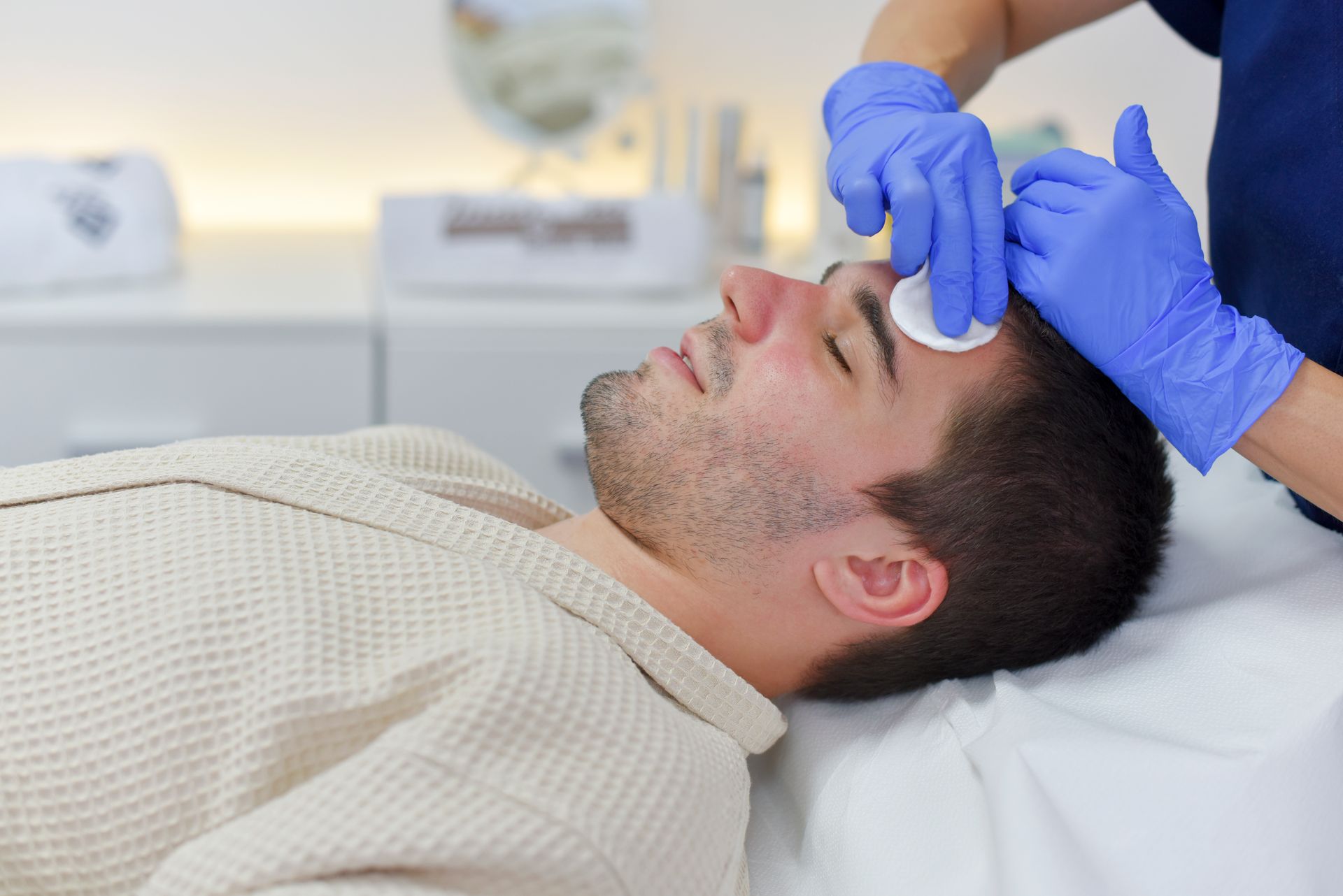 Man receiving a facial at a spa. A gloved esthetician cleans his forehead with cotton pads.