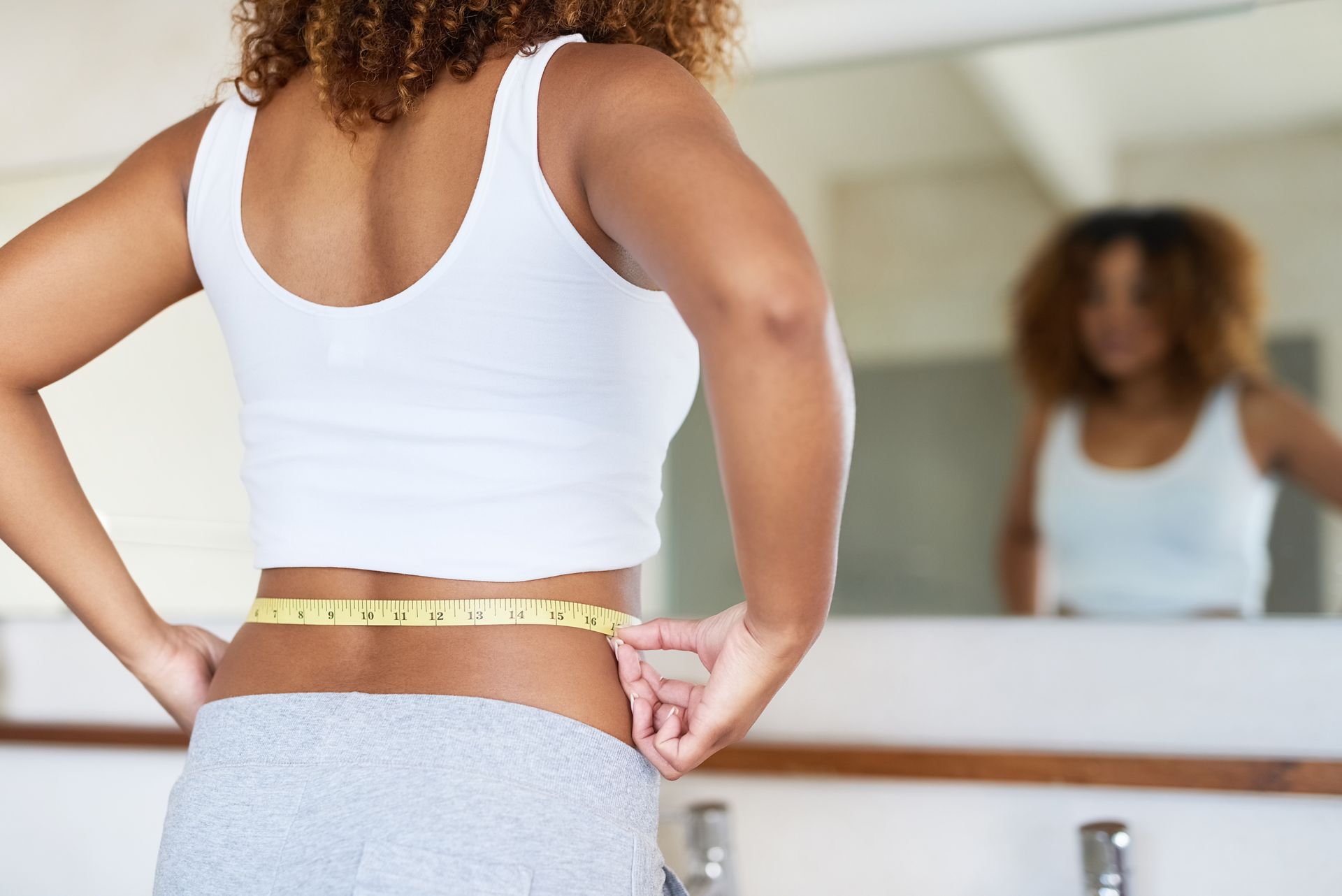 A person measures their waist with a yellow tape measure in front of a bathroom mirror.