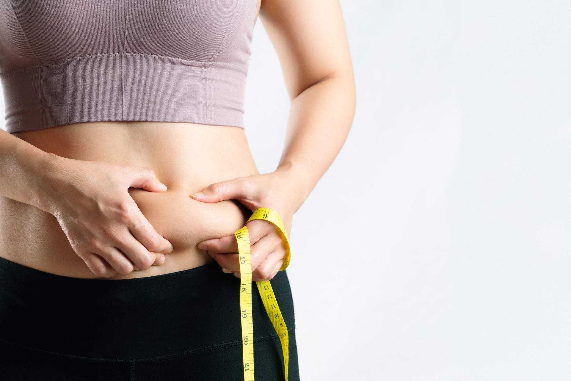 Woman in workout clothes pinching belly fat, measuring with a yellow tape.