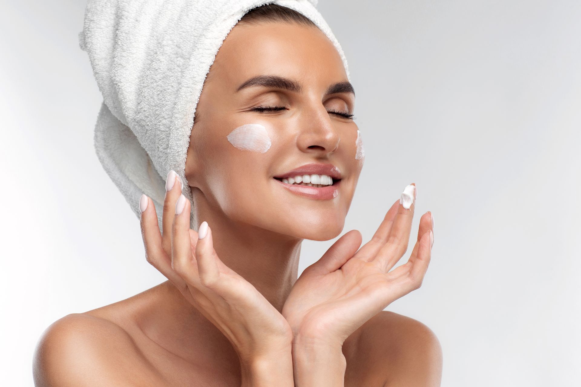 A woman with a towel wrapped around her head is smiling and applying cream to her face.