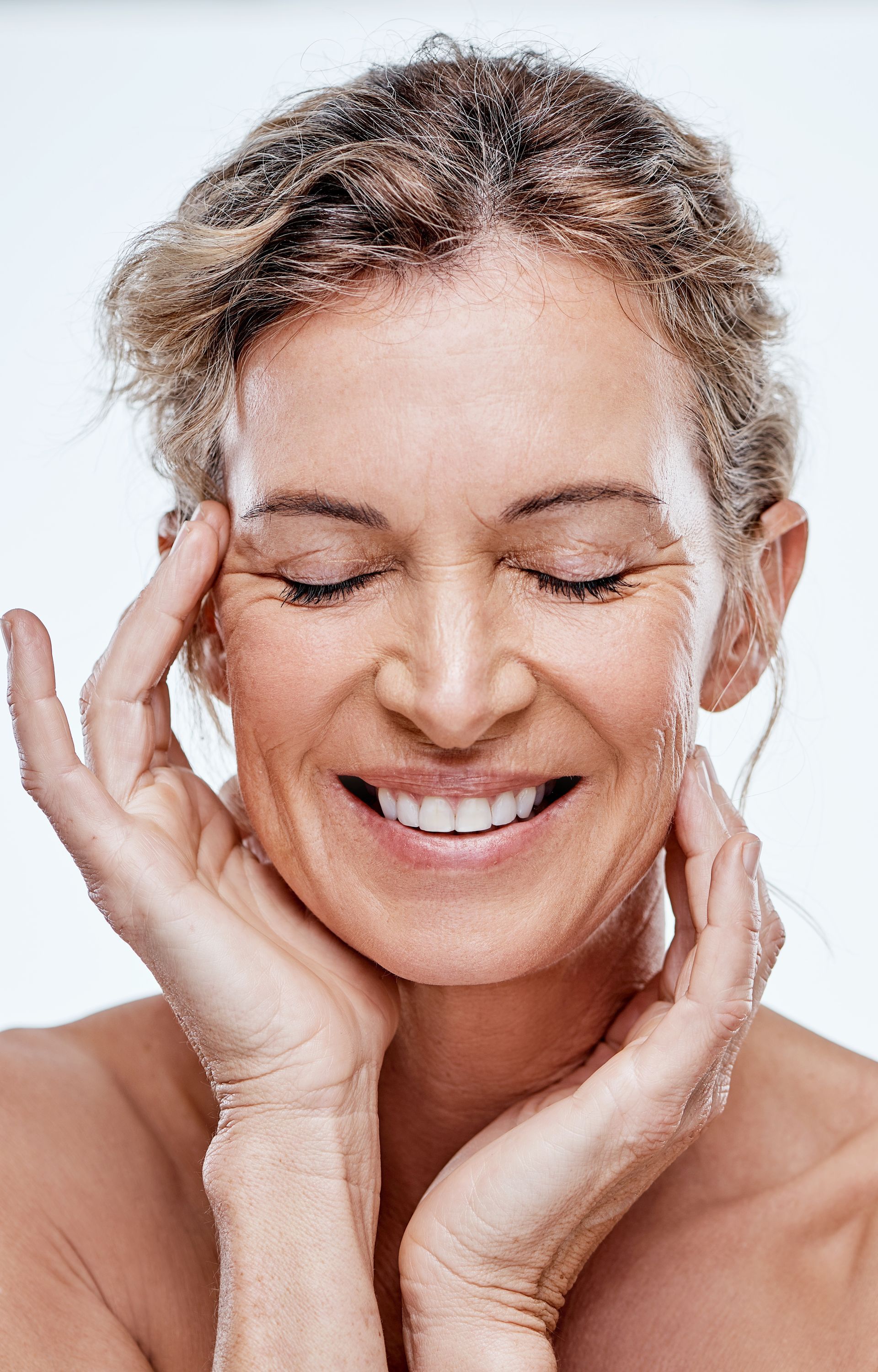 A person with eyes closed and a gentle smile, cradling their face with both hands against a plain white background.