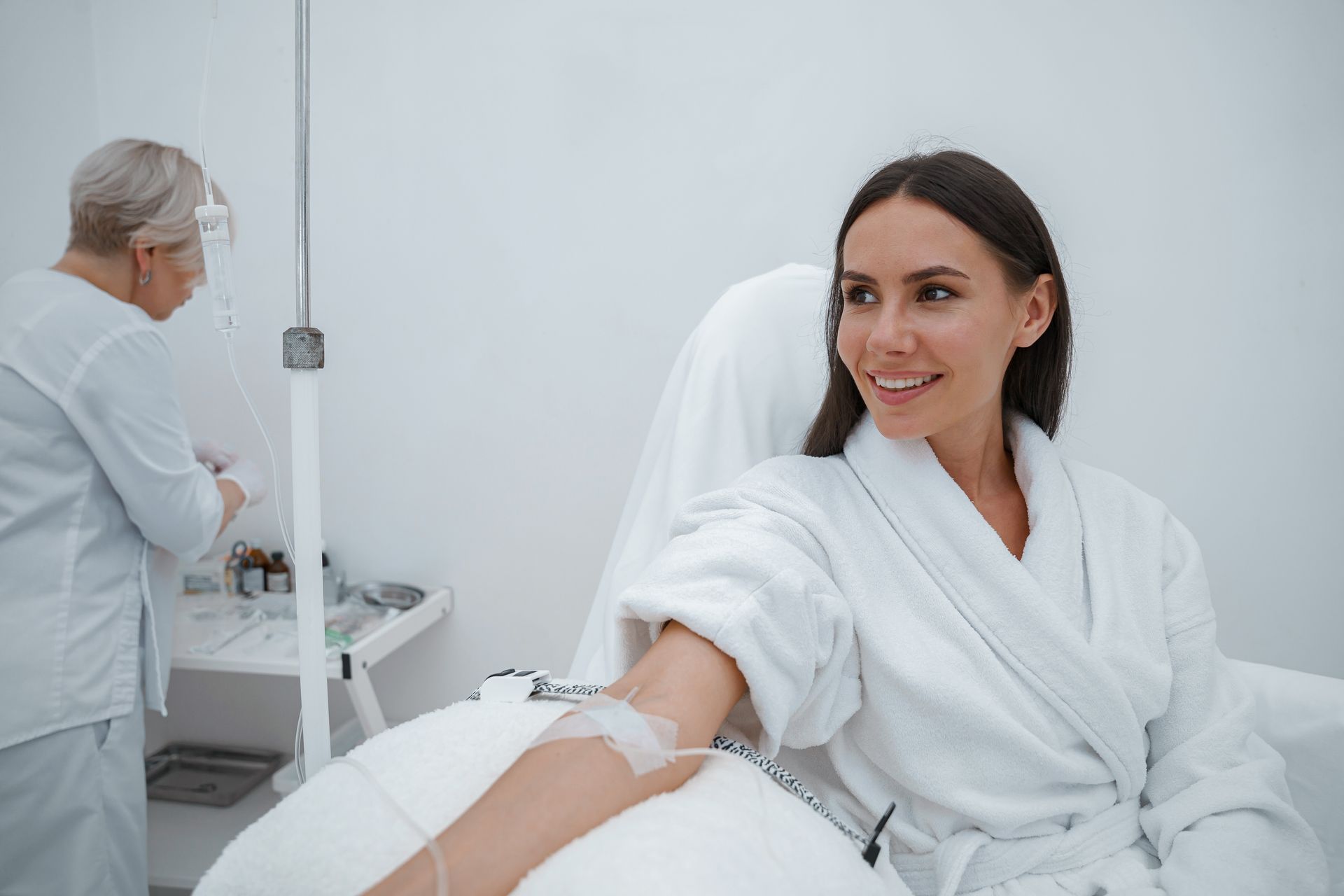 A person in a white robe receives an IV treatment while a medical professional prepares supplies in a clinical setting.