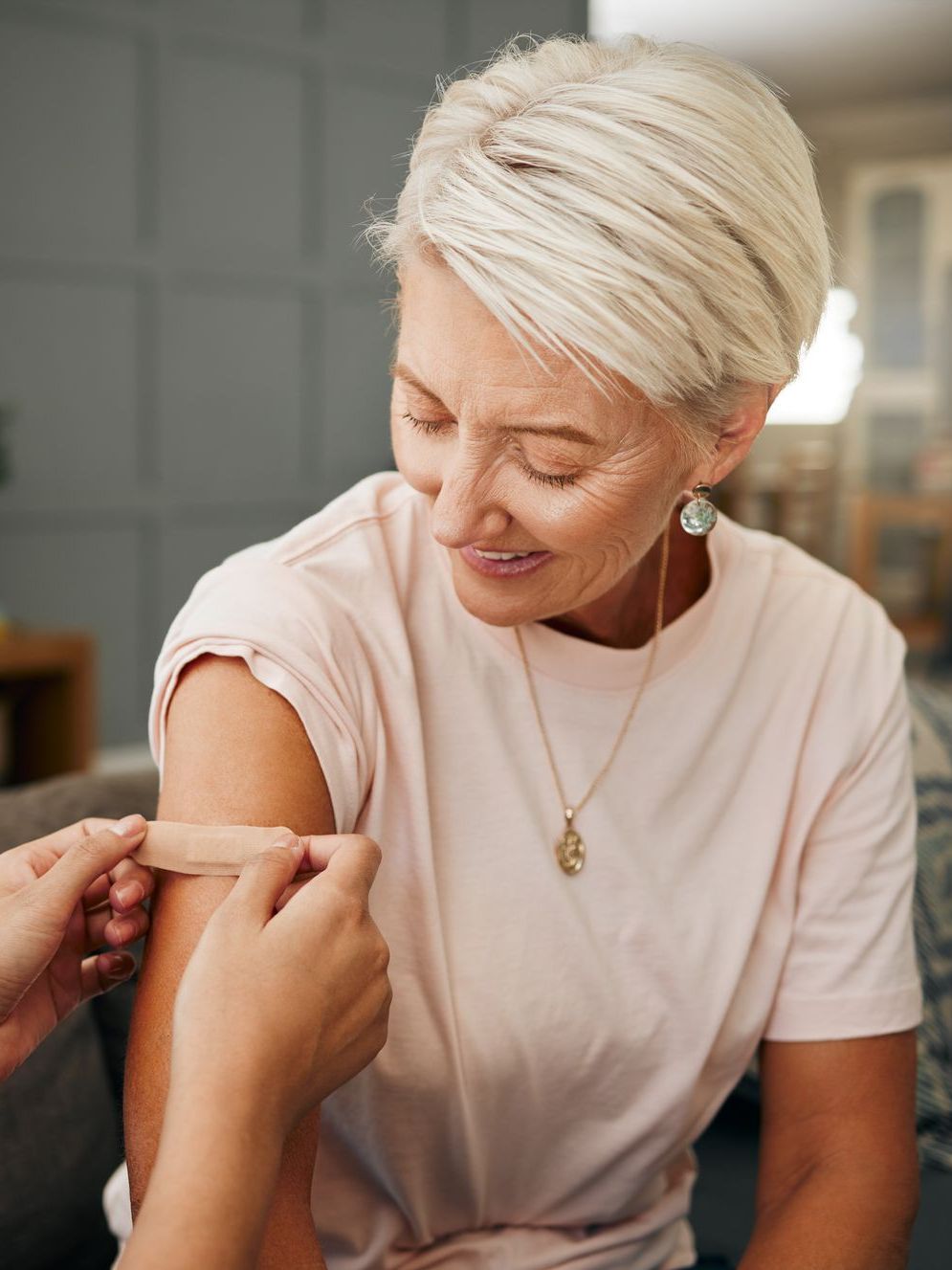 An older woman smiles as someone applies a bandage to her upper arm, likely after a shot, indoors.