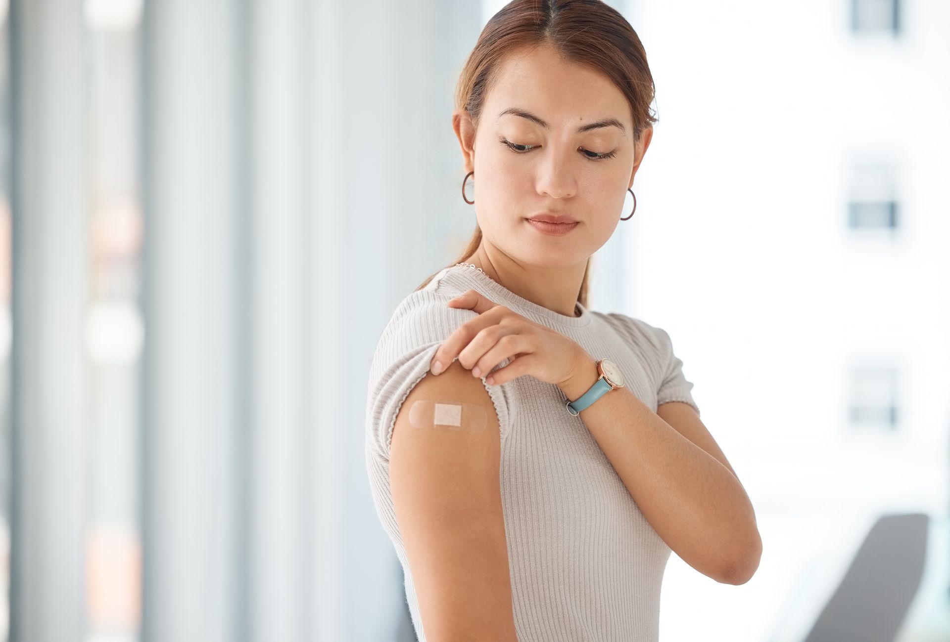 A woman with light skin looks at her arm where a bandage covers an injection site, likely after receiving a vaccine.