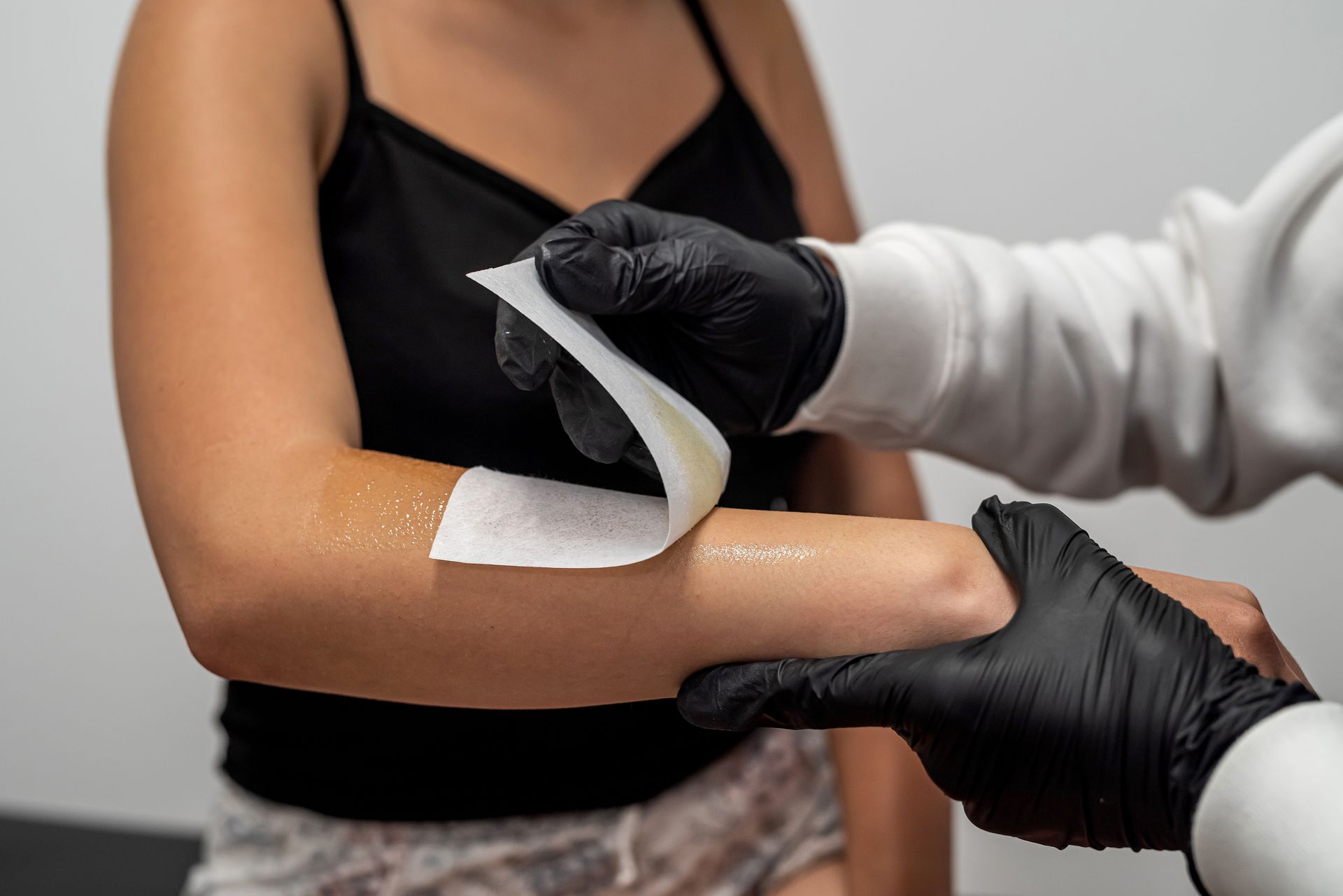 A person in black gloves applies a white wax strip to a forearm to remove hair in a salon setting.