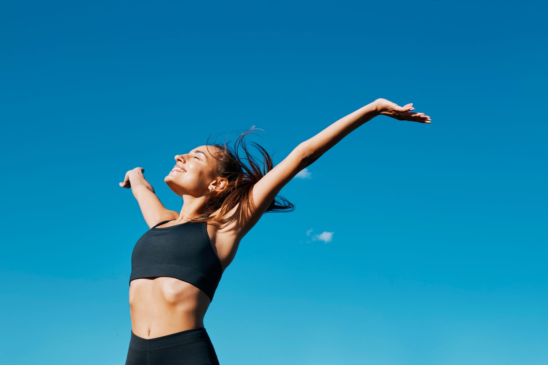 Woman in athletic wear with arms raised, smiling against a bright blue sky.
