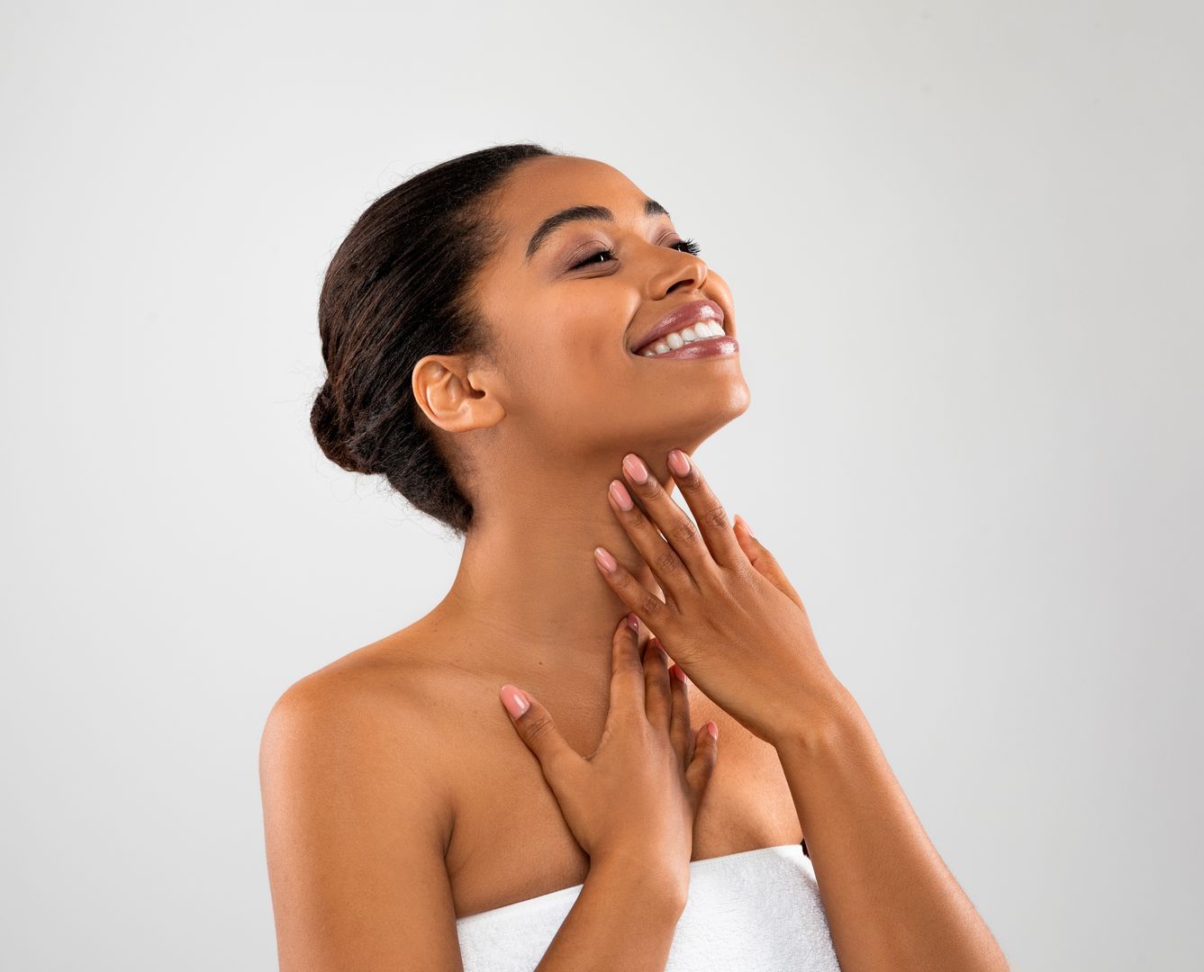 A woman wrapped in a towel is smiling and touching her neck.