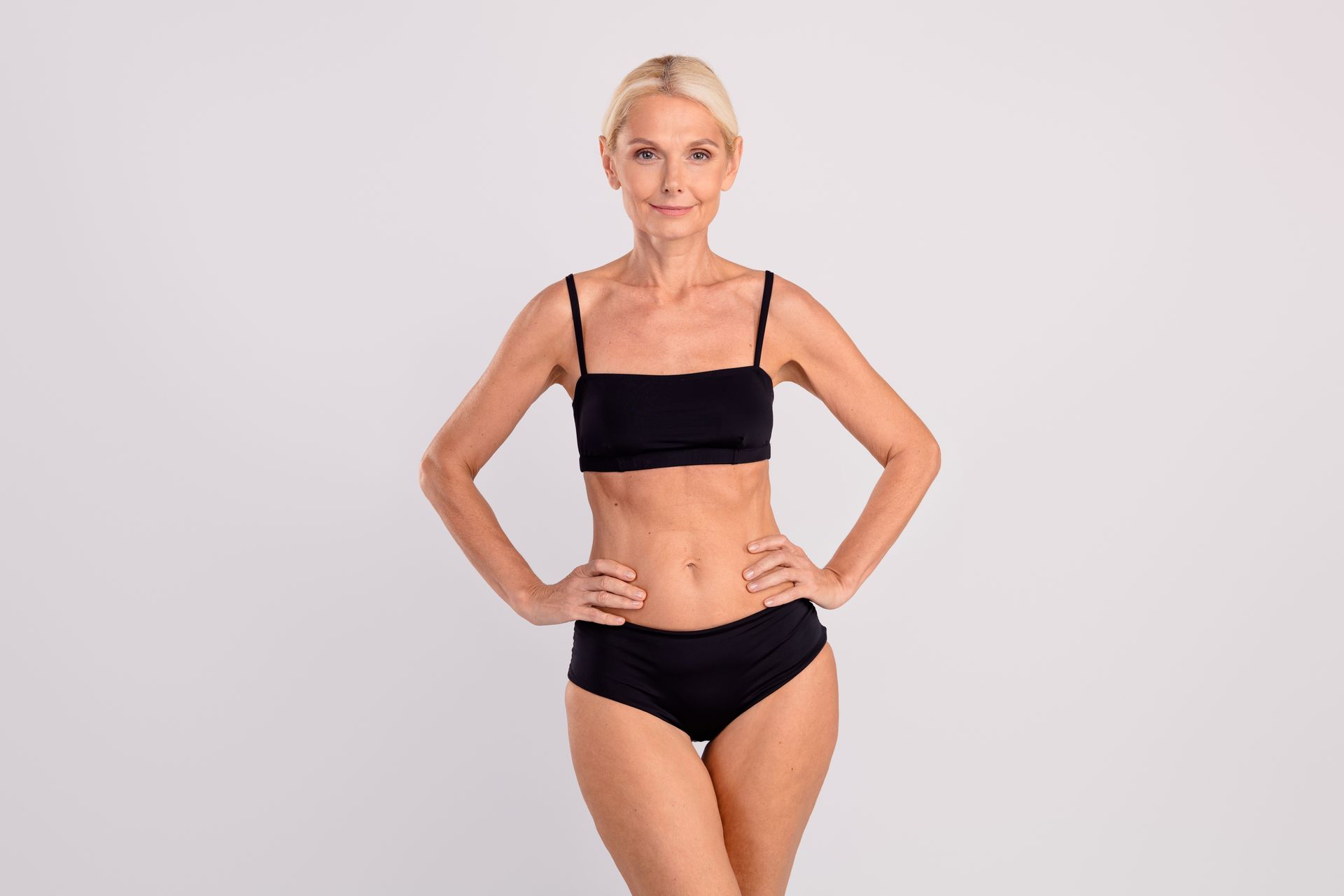 A person with blonde hair stands with hands on hips, wearing a black bikini against a plain white background.