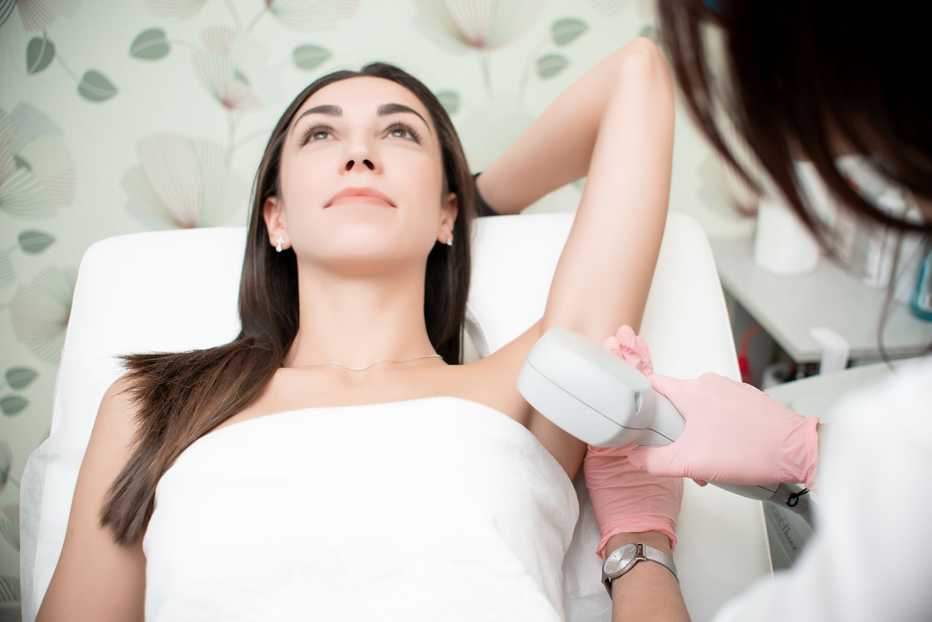 Woman receiving laser hair removal treatment on her underarm; light-skinned person holding device, wearing pink gloves.