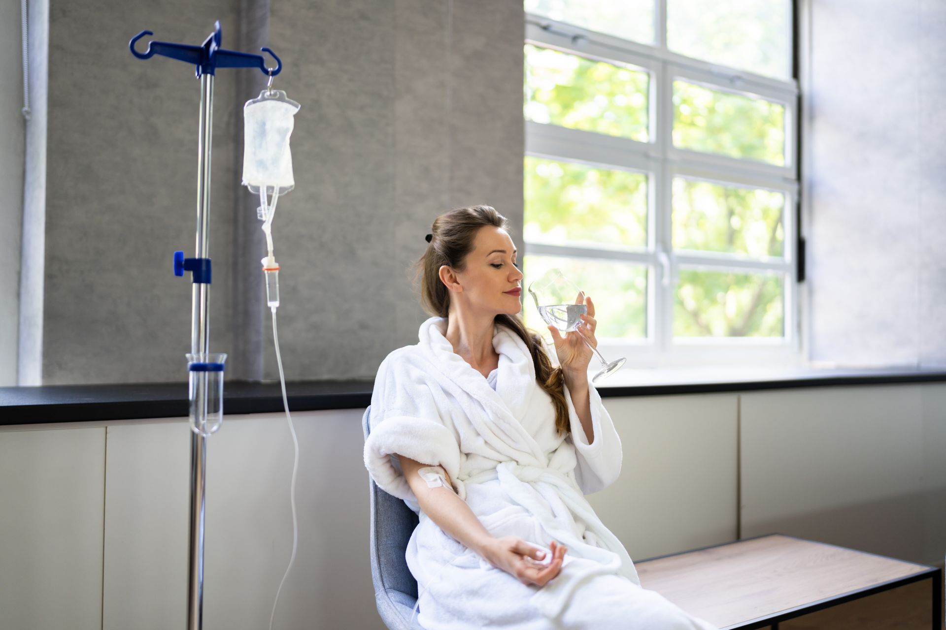 Woman in a bathrobe, receiving IV fluids, drinking water near a window.
