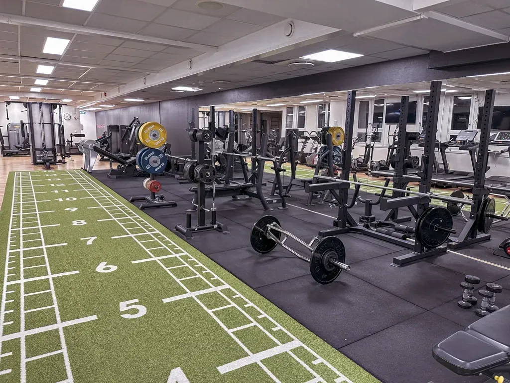 Et godt opplyst treningssenter med en lang grønn bane på gress, vektstativer og vektstenger på et svart gummigulv.