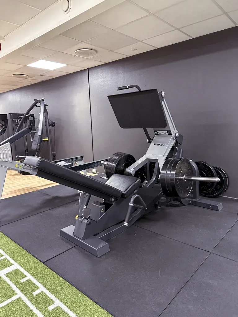 En beinpressmaskin lastet med vektskiver står på en svart gymmatte mot en mørkegrå vegg.