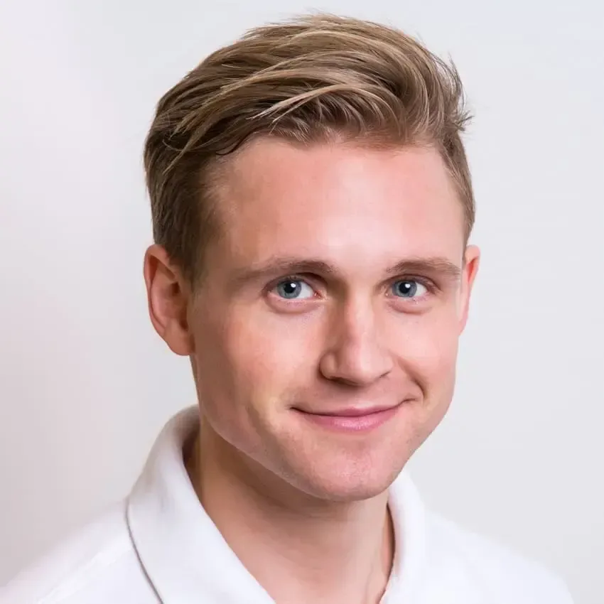 En mann med lyst hår og blå øyne smiler til kameraet, iført en hvit skjorte mot en lysegrå bakgrunn.