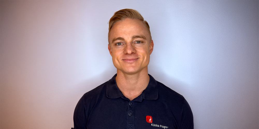 En person med blondt hår iført en marineblå poloskjorte med rød logo, smilende mot en lysegrå bakgrunn.