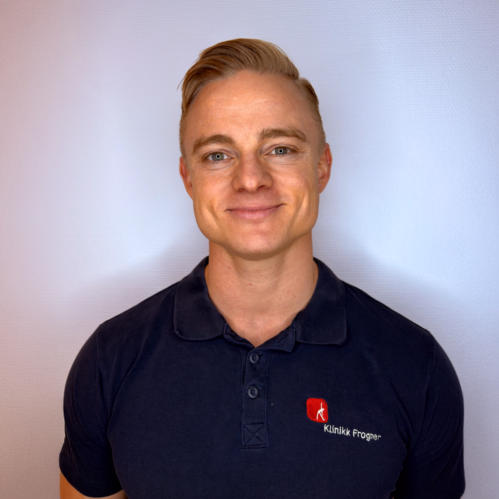 En person med blondt hår iført en marineblå poloskjorte med rød logo, smilende mot en lysegrå bakgrunn.