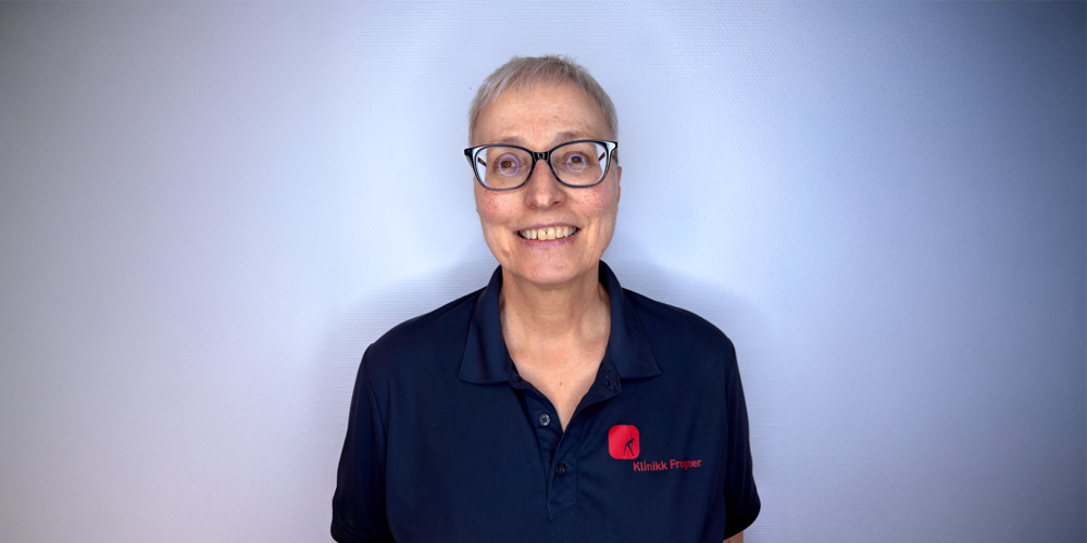 En person med briller og en marineblå poloskjorte med en liten rød logo, smilende mot en lyseblå bakgrunn.