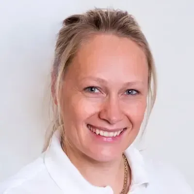 En kvinne med blondt hår i hvit skjorte smiler til kameraet mot en hvit bakgrunn.