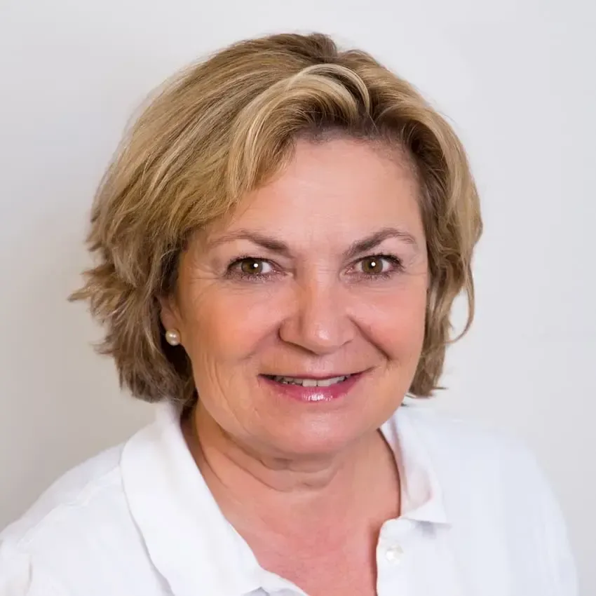 Kvinne med kort blondt hår, smilende, iført hvit skjorte, mot en hvit bakgrunn.