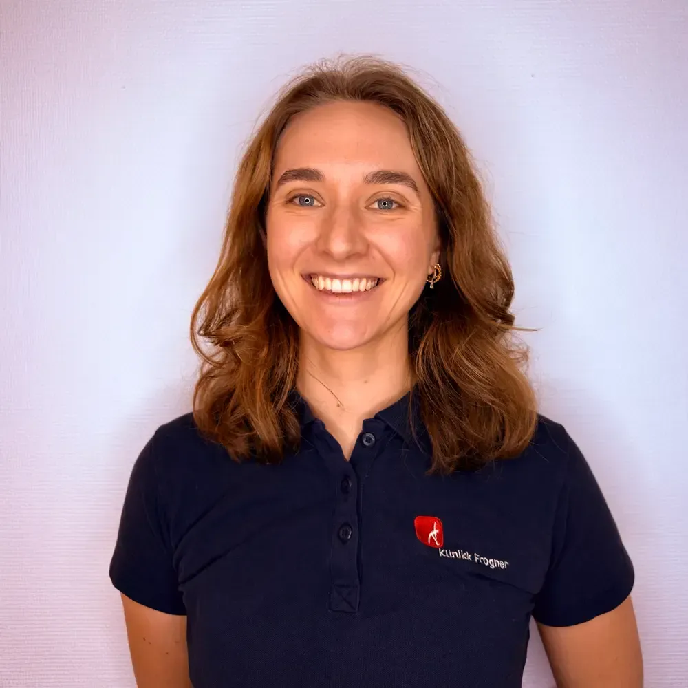 En smilende person med skulderlangt brunt hår har på seg en mørkeblå poloskjorte med en liten rød logo på brystet.