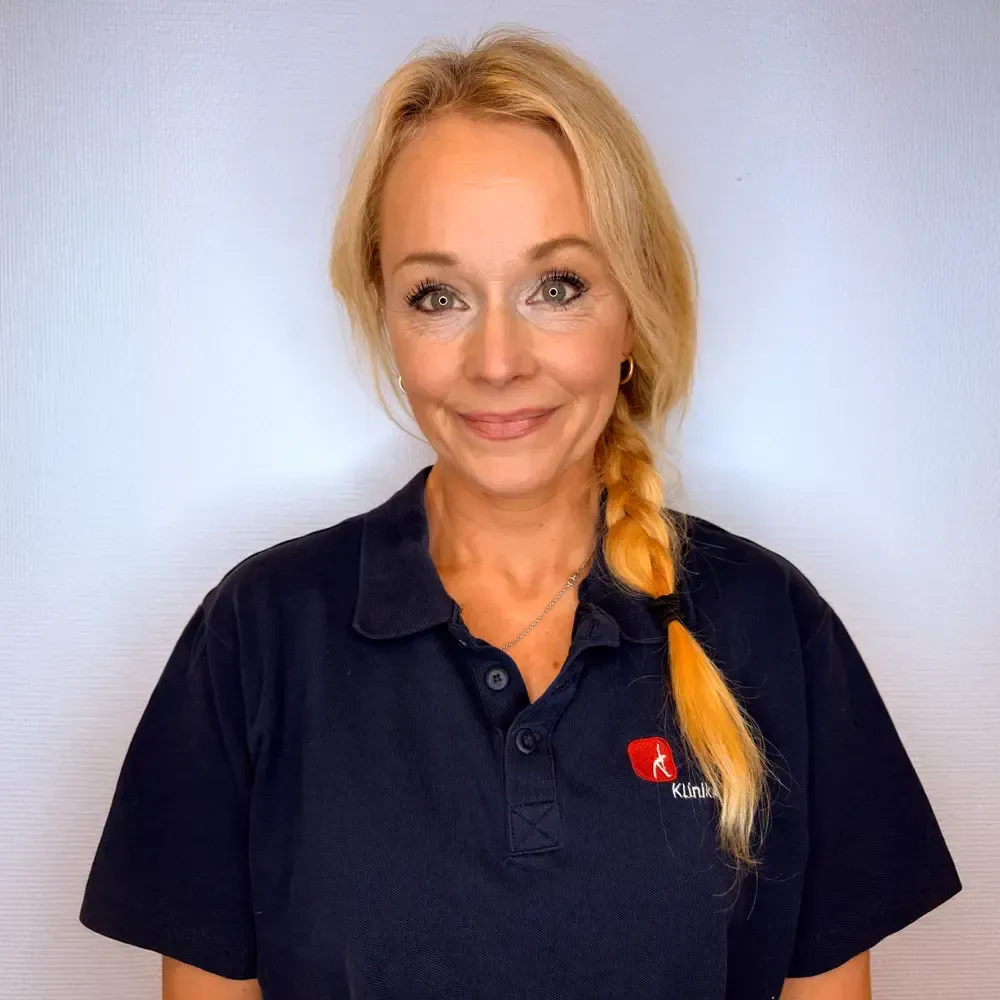 Kvinne med blondt hår i en lav hestehale iført en mørkeblå poloskjorte, smilende, mot en hvit bakgrunn.