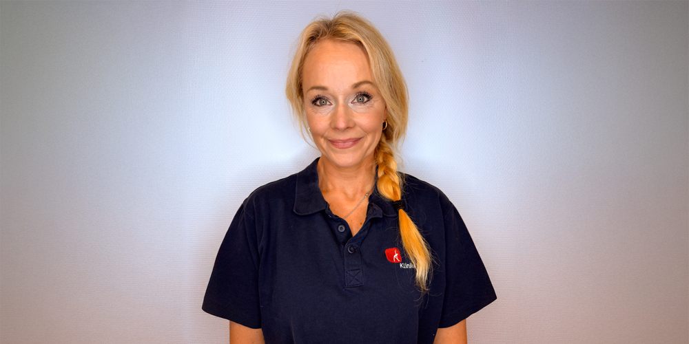 Kvinne med blondt hår i en lav hestehale iført en mørkeblå poloskjorte, smilende, mot en hvit bakgrunn.