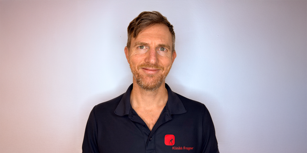 En smilende person iført en mørkeblå poloskjorte med en liten rød firkantet logo på brystet, stående inntil en enkel vegg.