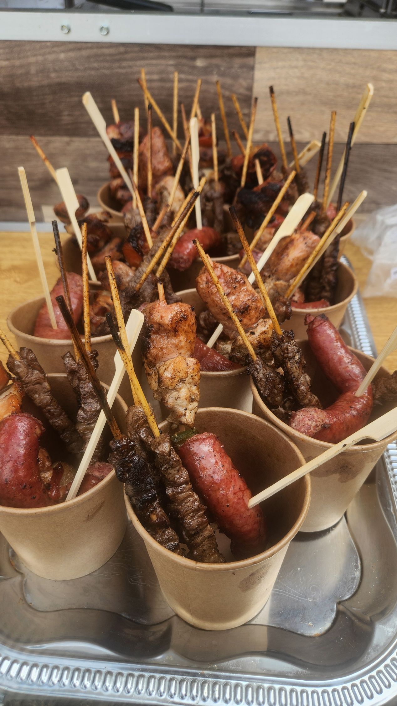Vassoio di salsicce arrosto infilzate su spiedini e servite in bicchierini di carta, condite con salsa e spezie.