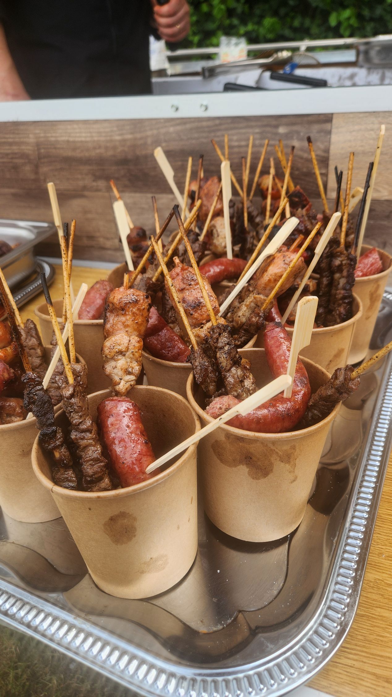 Spiedini di carne grigliata assortiti in bicchieri di carta su un vassoio presso una bancarella di cibo all'aperto.