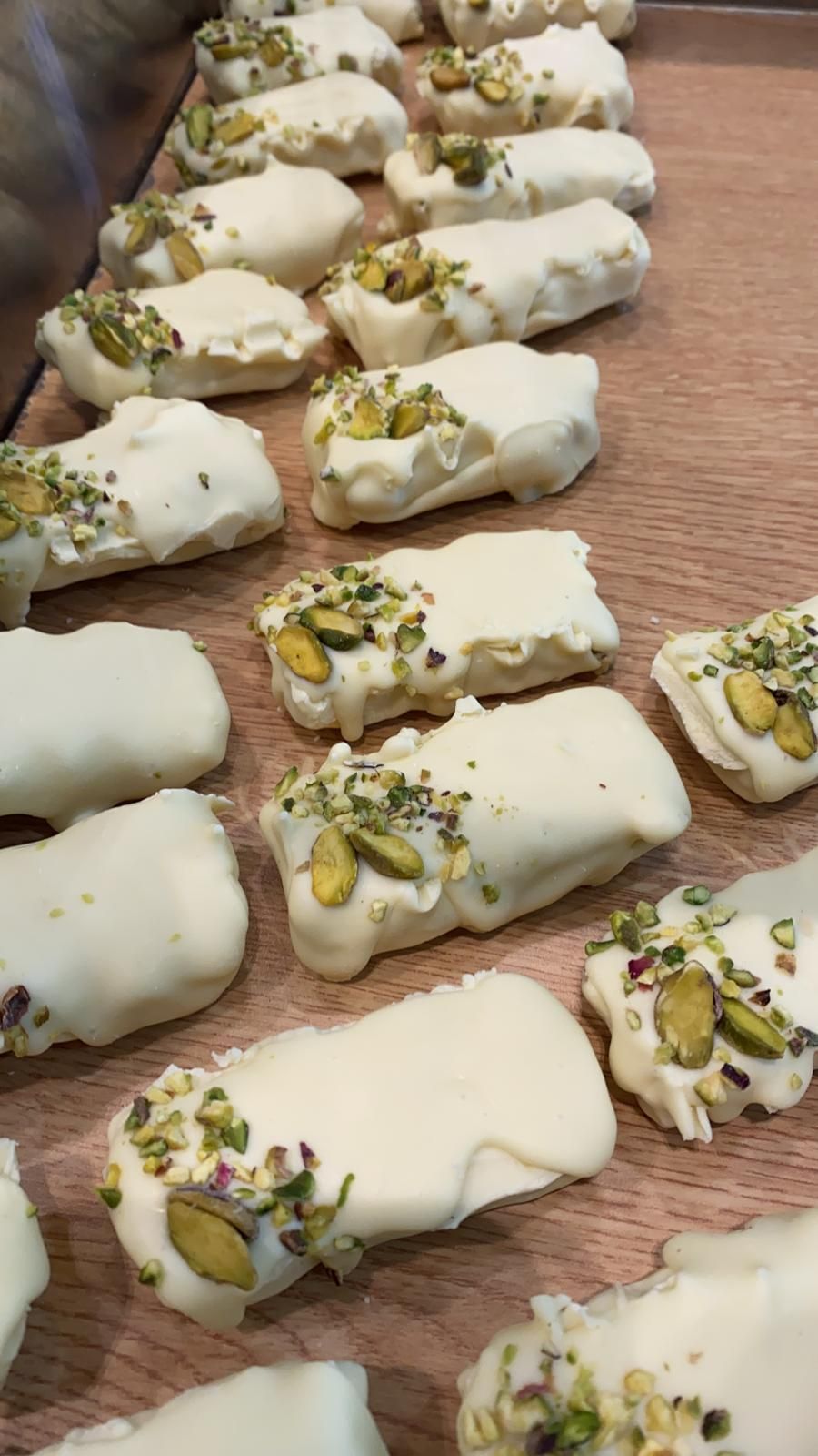 File di dolcetti ricoperti di cioccolato bianco e cosparsi di pistacchi tritati, disposti su un vassoio di legno.
