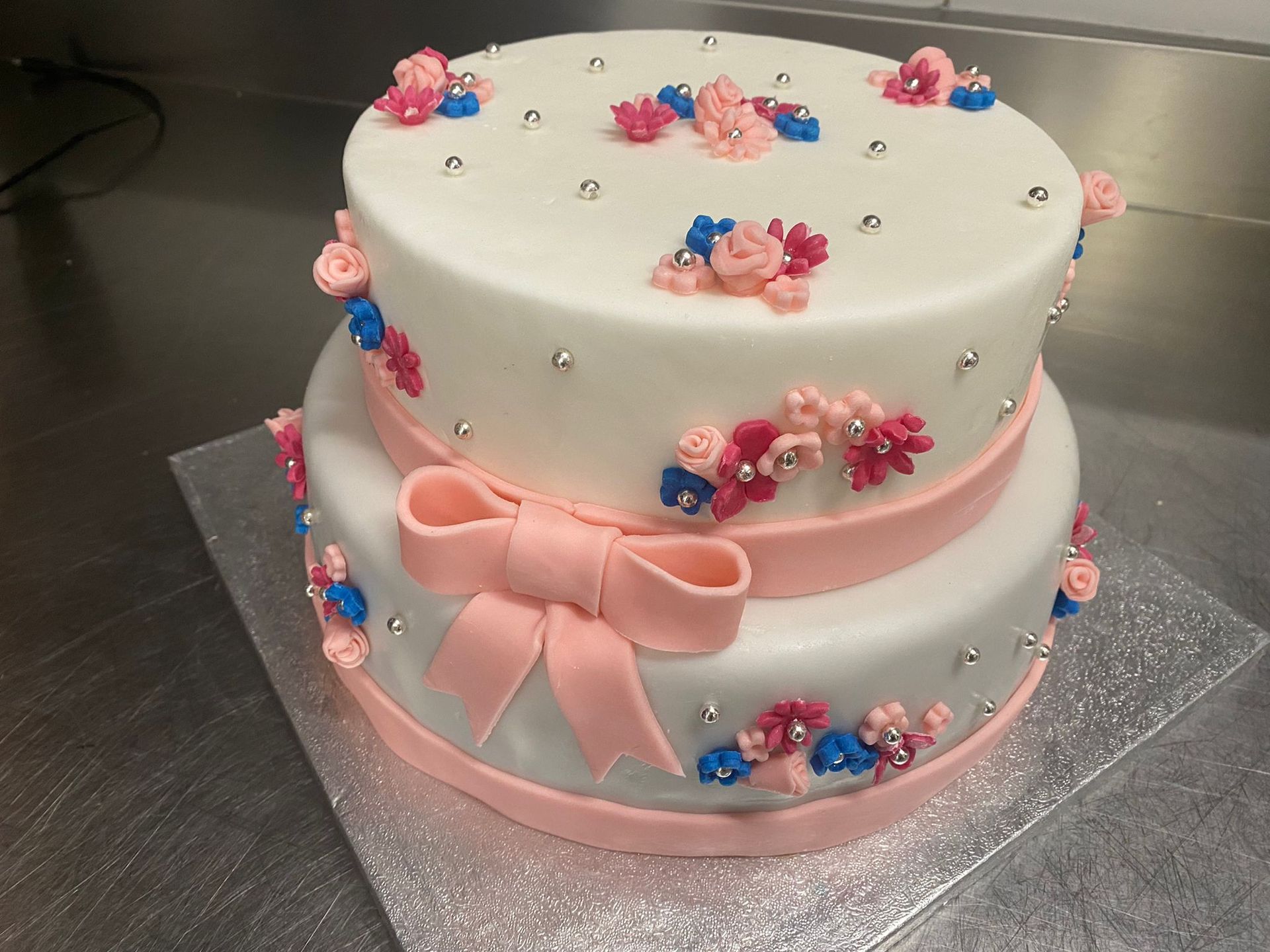 Torta bianca a due piani con fiocco di nastro rosa e decorazioni floreali su un vassoio d'argento.