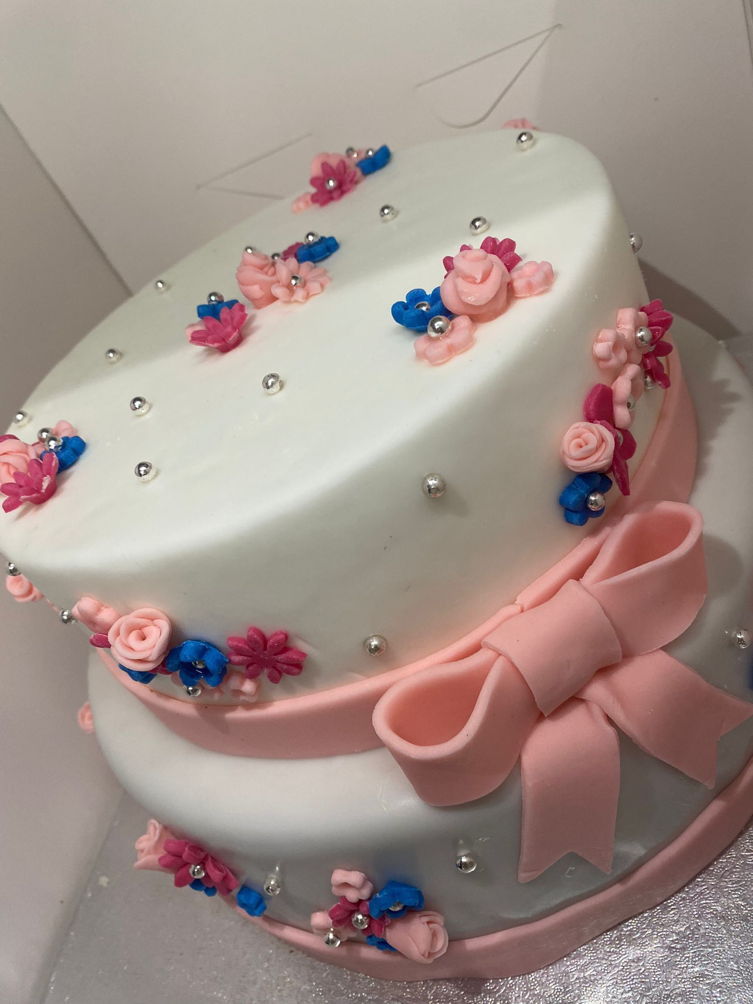 Torta a più piani bianca con nastro rosa, fiori e perline argentate su una base color pastello.