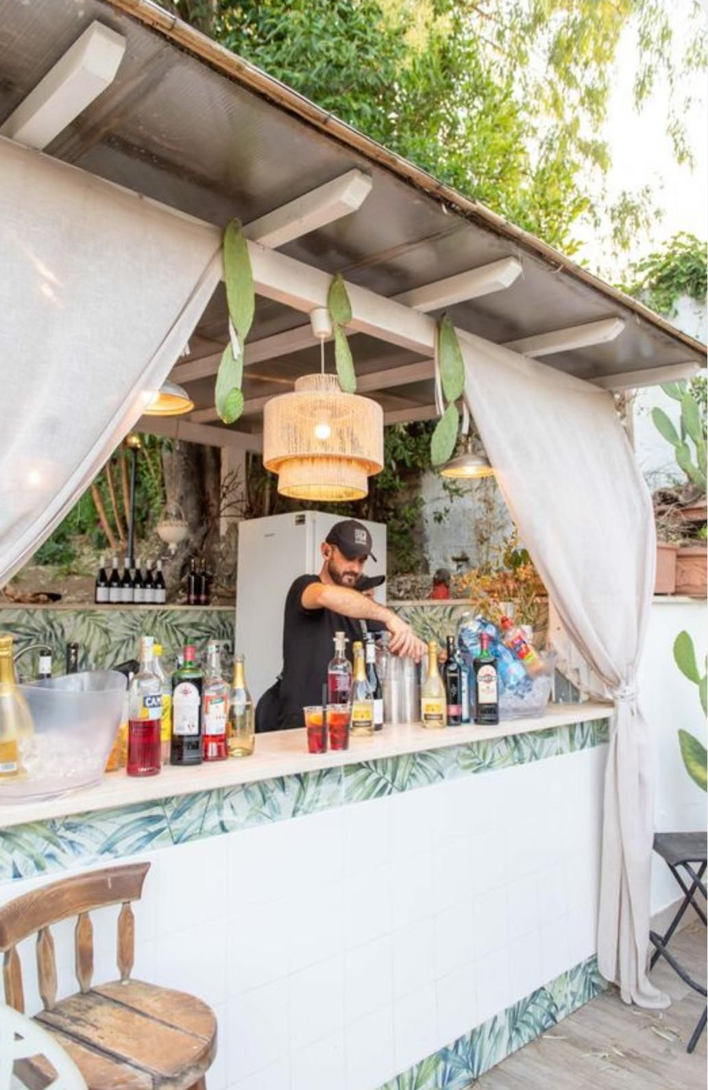 Cocktail bar all'aperto con bottiglie sul bancone e un barista che serve i clienti sotto una tettoia di paglia.