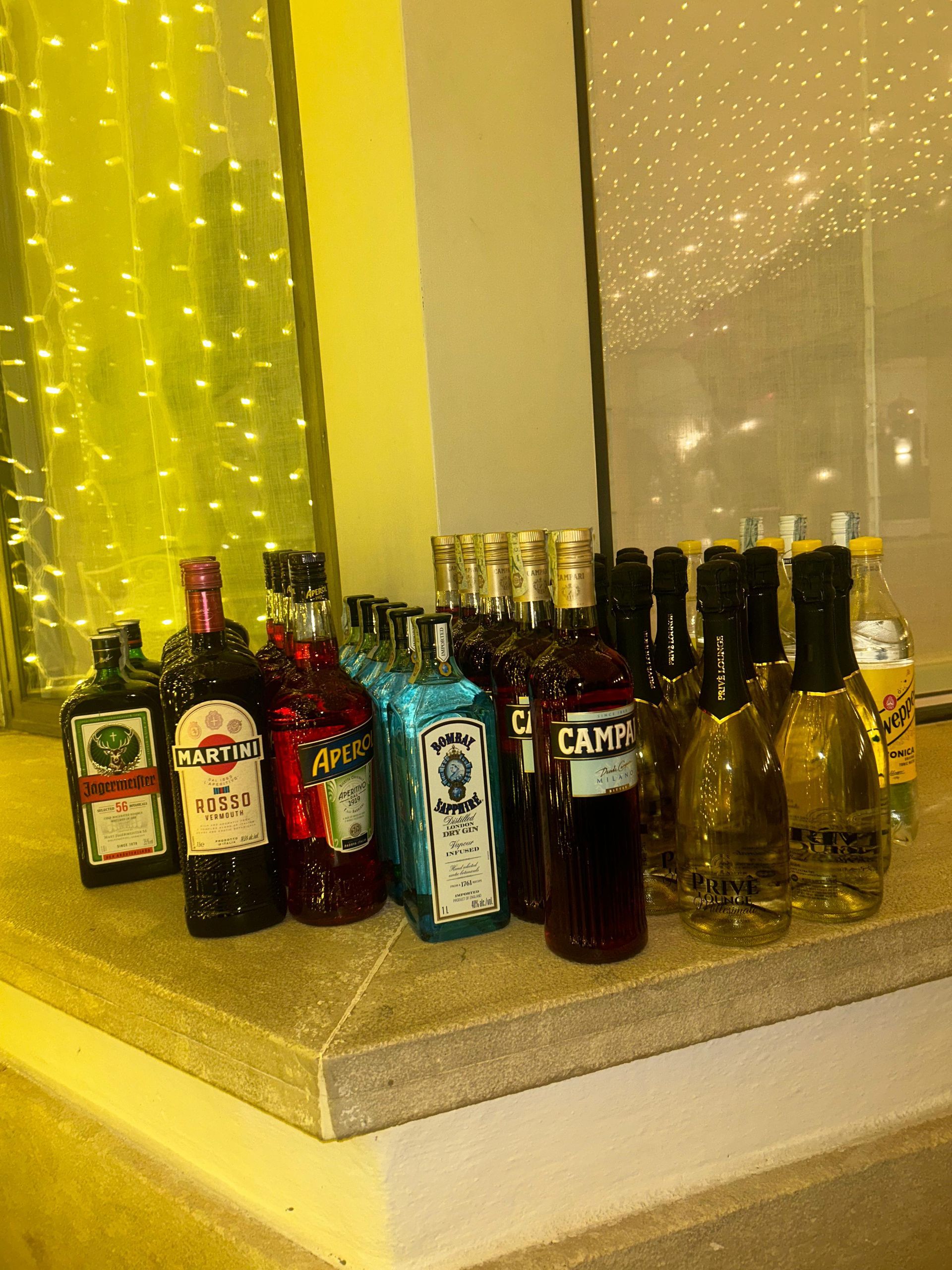 Diverse bottiglie di liquore su un ripiano accanto a una finestra illuminata di giallo e con le tende.