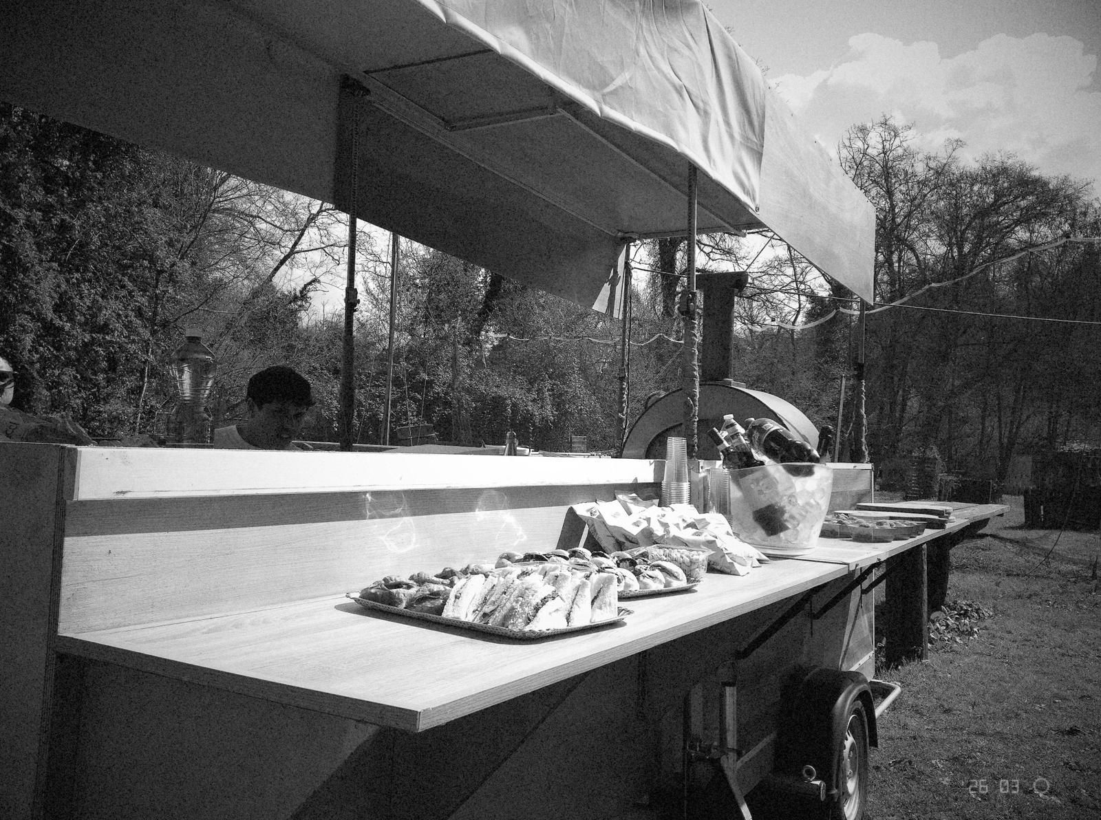 Tavolo da buffet all'aperto sotto una tettoia, con vassoi di cibo e distributore di bevande in bianco e nero.