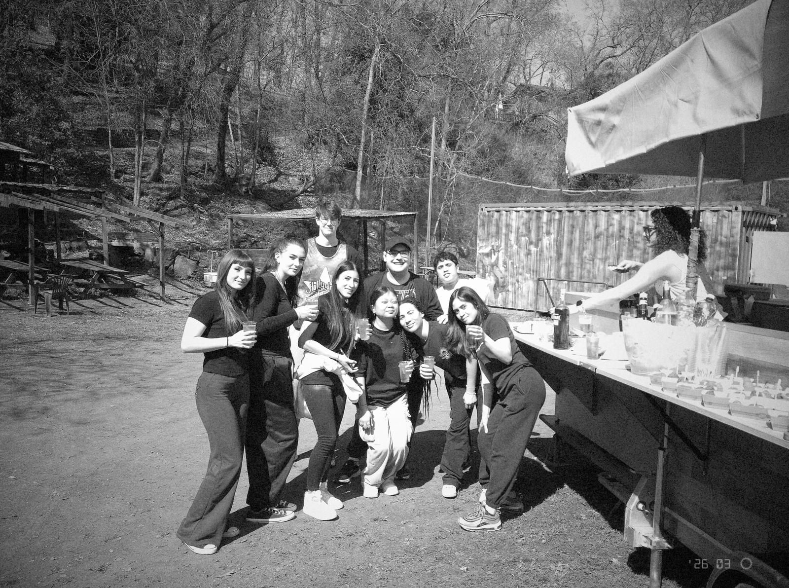 Gruppo di persone in posa all'aperto accanto a una bancarella di cibo, con alberi e una recinzione sullo sfondo.