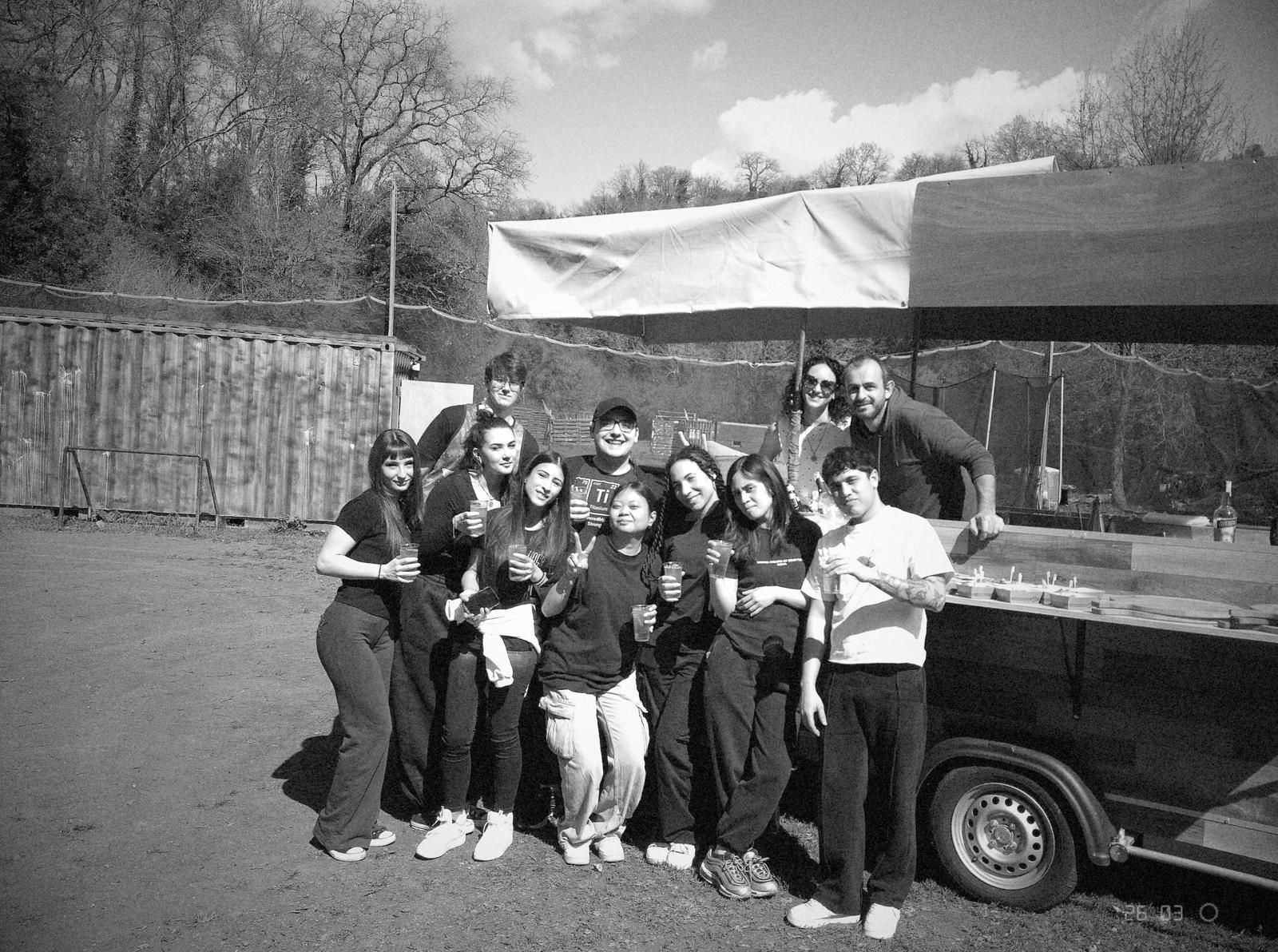 Gruppo di persone in posa accanto a un'auto con rimorchio parcheggiata all'aperto.