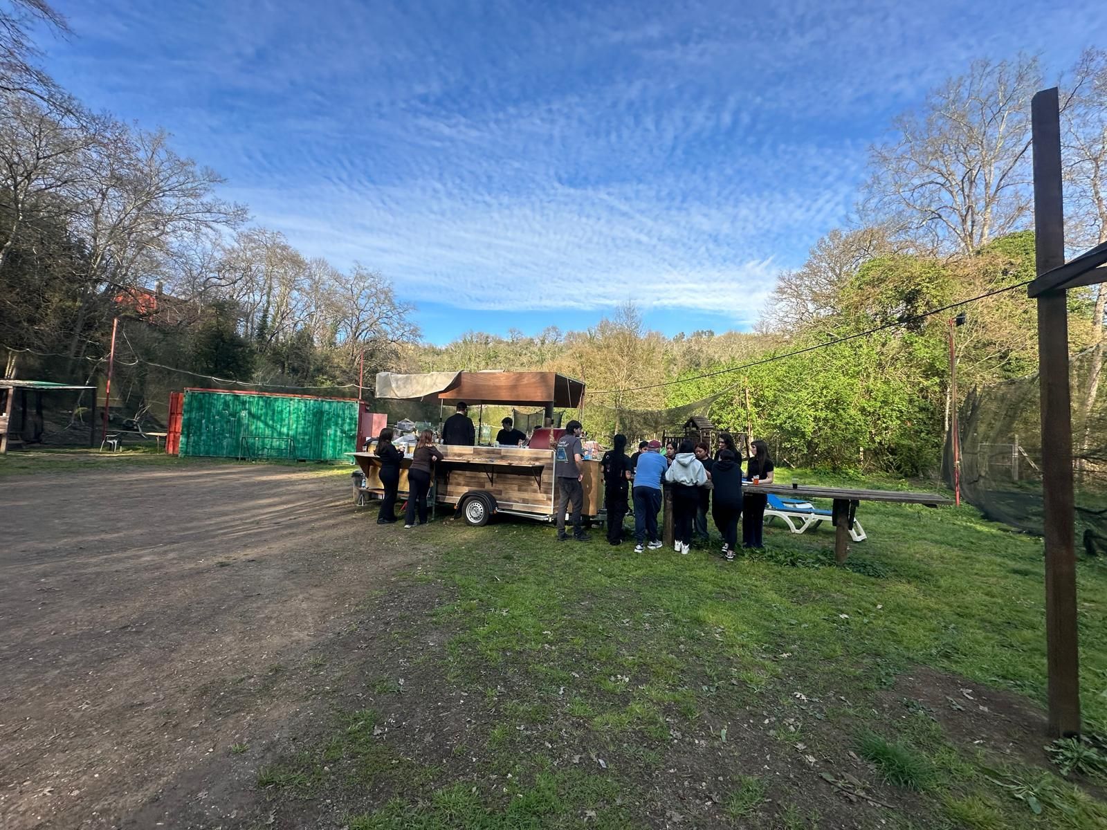 In una giornata di sole, diverse persone si sono radunate attorno a un rustico chiosco di cibo all'aperto in un parco.