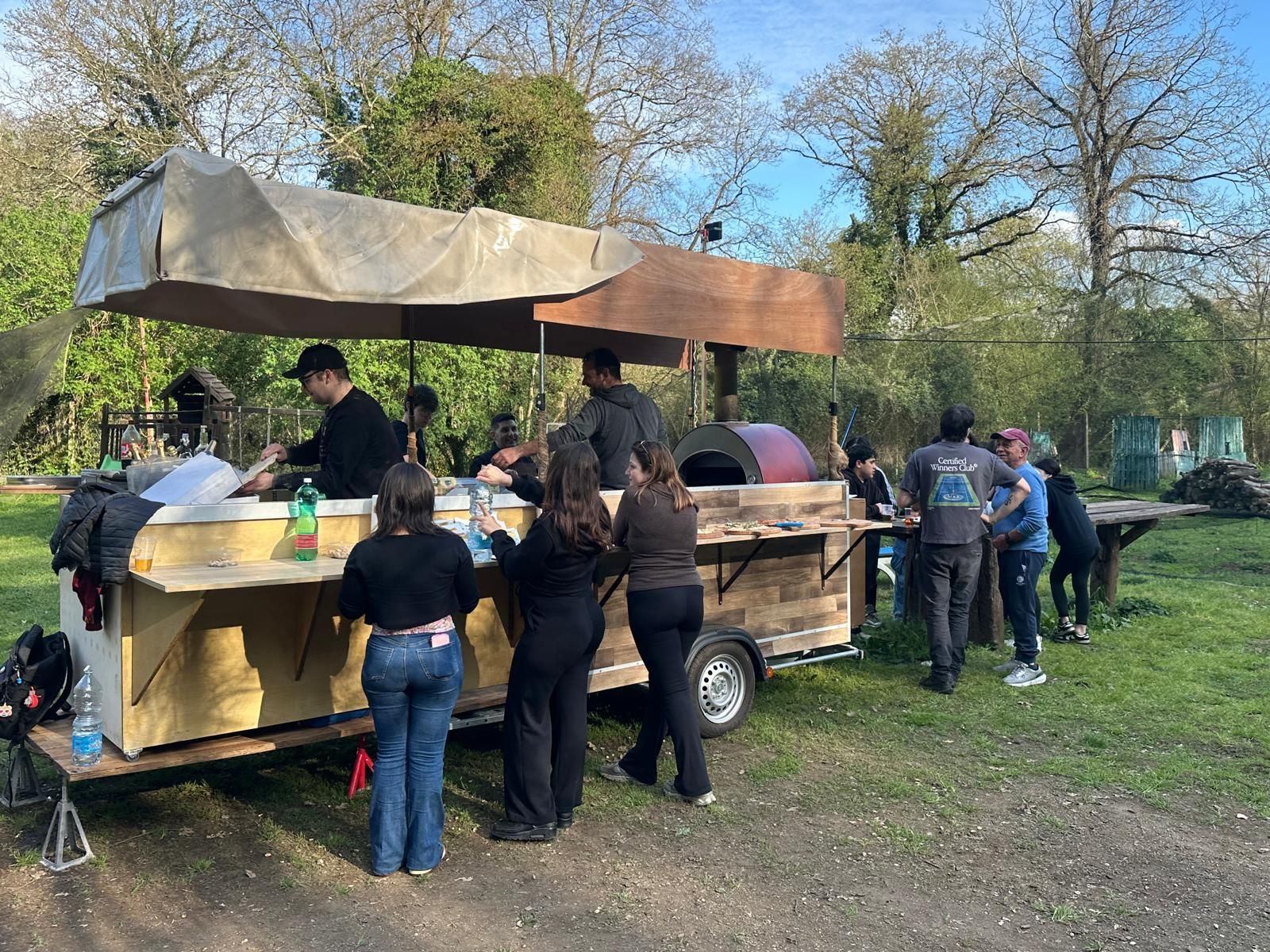 In un parco, diverse persone si sono radunate attorno a una bancarella di cibo all'aperto, sotto una tettoia.