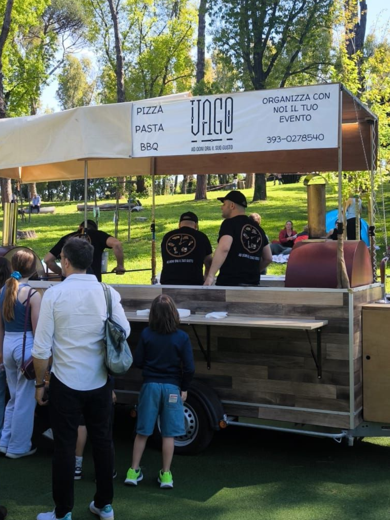 Bancarella di cibo all'aperto sotto una tettoia, con persone che ordinano durante una fiera in un parco.
