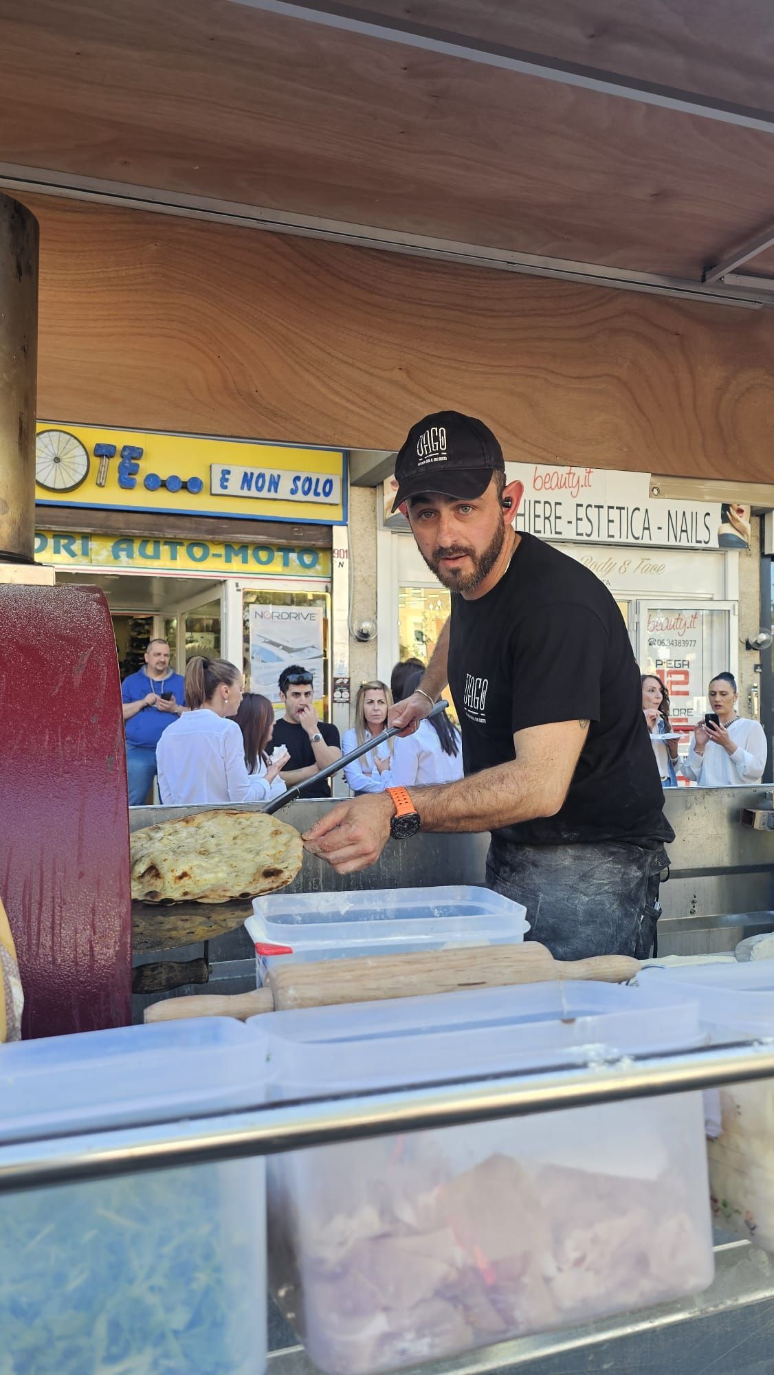 Un venditore ambulante griglia carne in una bancarella all'aperto, indossando un berretto e una maglietta neri.