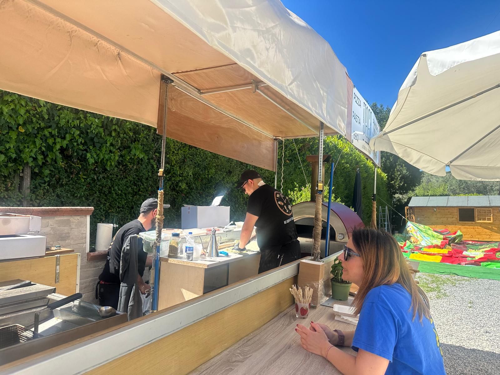 Chiosco di cibo all'aperto sotto gli ombrelloni, con il personale che serve i prodotti a un cliente seduto in un patio soleggiato.