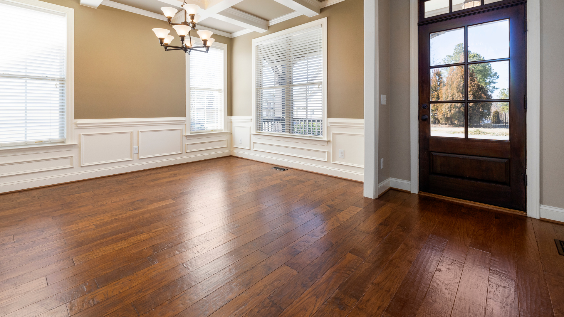 Empty room with hardwood floors, windows, and a dark wooden door. The walls are neutral colors.