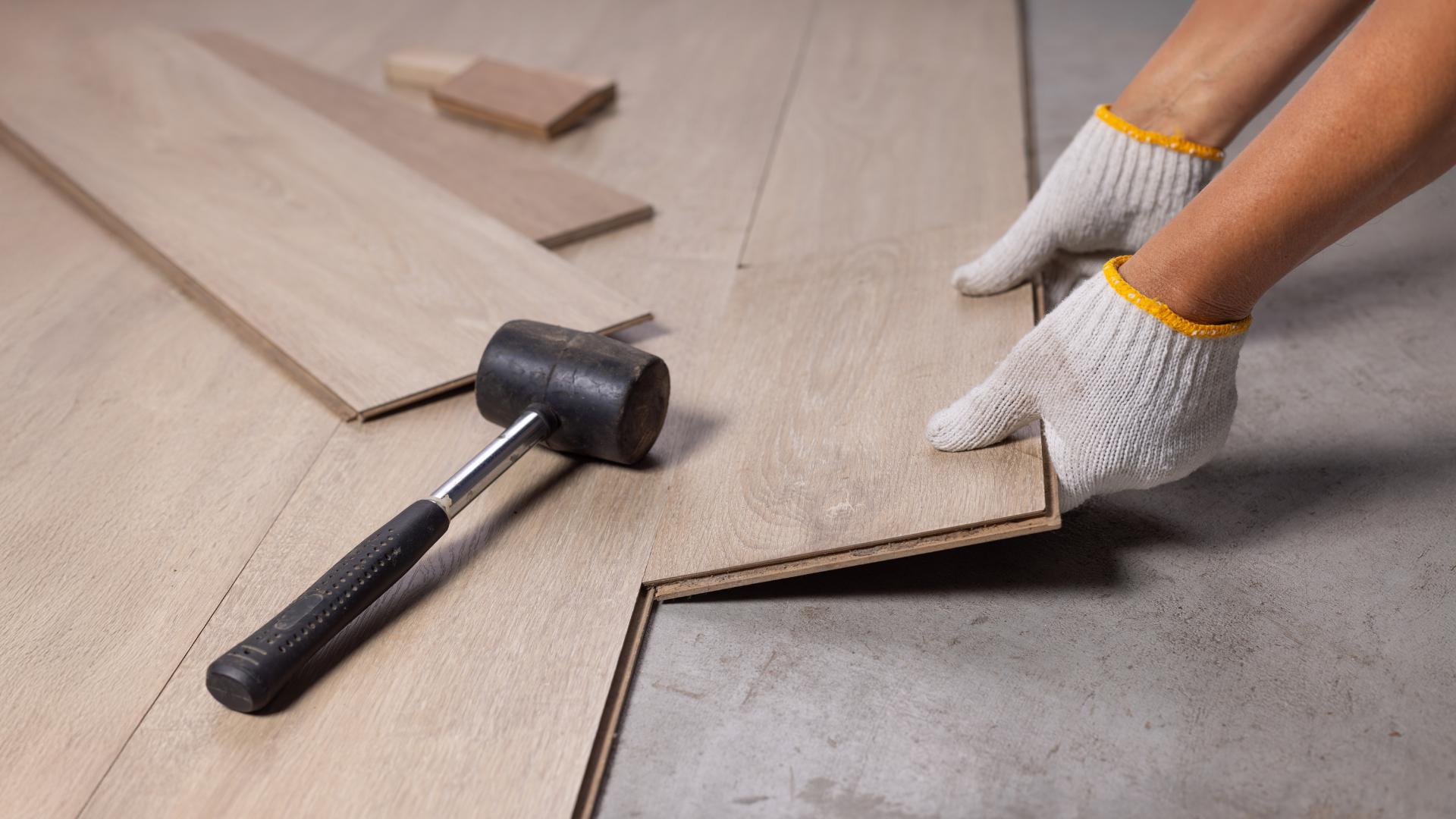 Hands wearing gloves installing wood flooring, using a hammer.